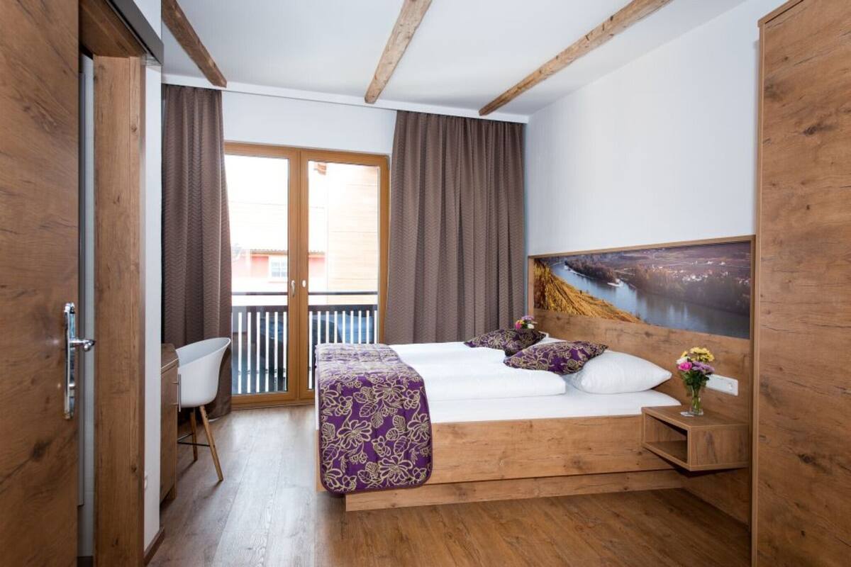 Une chambre avec un lit en bois, un bureau et une chaise, un balcon avec des rideaux et un tableau accroché au mur.