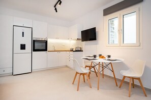 A modern kitchen with white cabinets, a built-in oven, a dining table with chairs, and a window with blinds. - Naxos Mini Cube (Naxos)