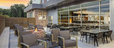 An outdoor seating area with wicker furniture and a fire feature, adjacent to a building with large windows and a brick facade.