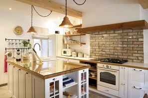 A kitchen with a central island, wooden beams, a brick wall, and white cabinetry. - Pri Stefacovih – Authentic house - Sauna and Hot tub (Žerovnica)