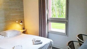 Et hotelværelse med en seng, et natbord med lampe, en stol og et vindue med udsigt til træer.