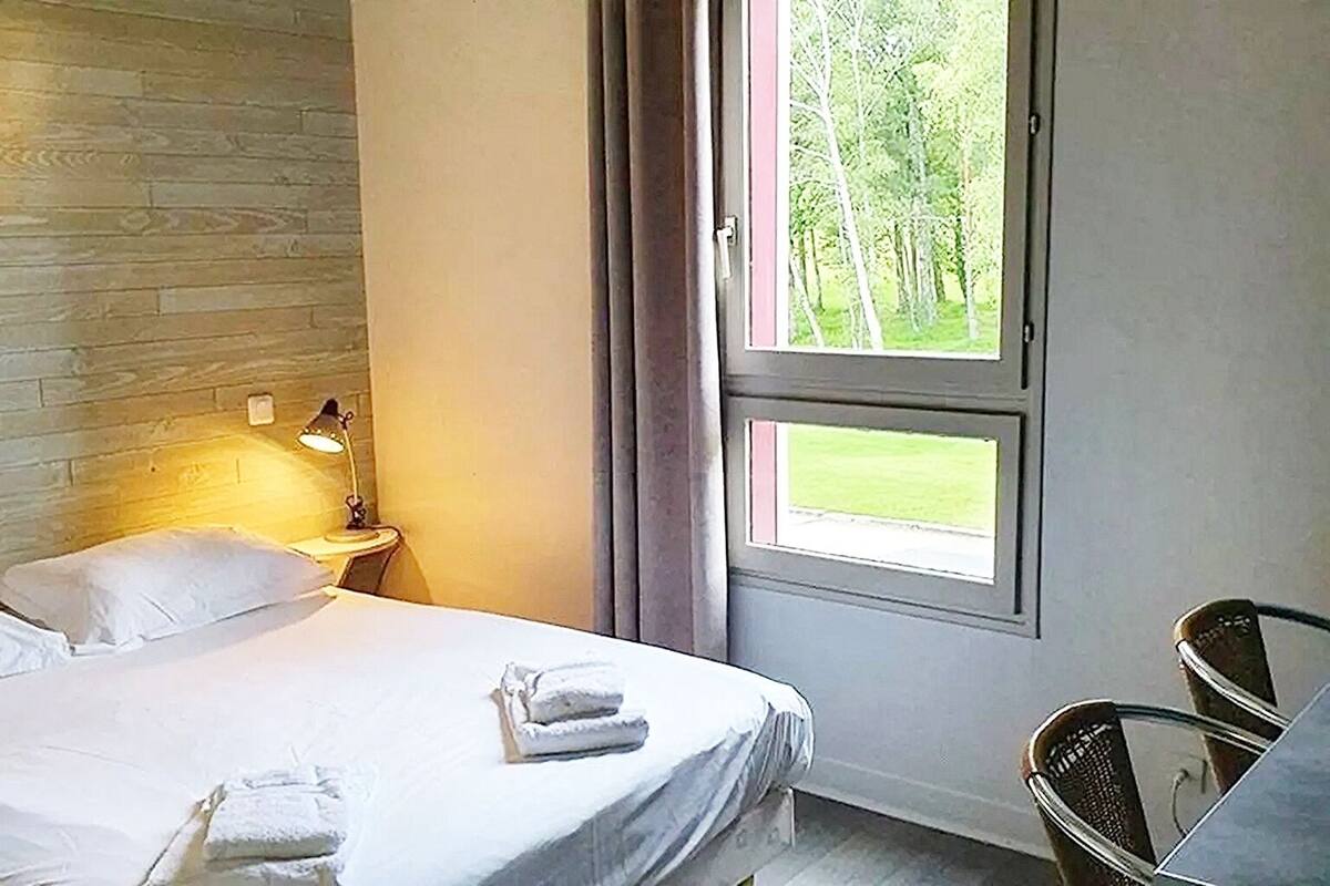 Una camera d'albergo con un letto, un comodino con una lampada, una sedia e una finestra vista alberi.