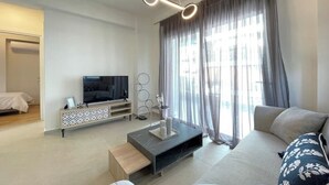 Ein modernes Wohnzimmer mit Flachbildfernseher, einer Couch, einem Couchtisch und einem großen Fenster mit Vorhängen.
