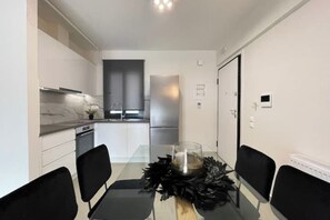 Et moderne køkken med hvide skabe, et rustfrit stål køleskab og et spiseområde med et glasbord og sorte stole.