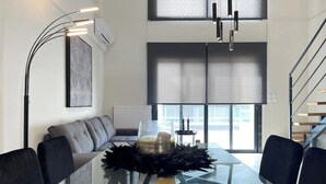 Ein modernes Wohnzimmer mit einem Glastisch, schwarzen Sesseln, einem grauen Sofa, einem schwarzen Dekoartikel und einem großen Fenster mit Jalousien.