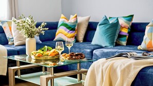 Een woonkamer met een blauwe bank, kleurrijke kussens, een glazen salontafel met fruit, glazen en een boek.