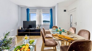 Een moderne woonkamer met een flatscreen televisie, een eettafel met stoelen, een glazen salontafel met fruit, en uitzicht op het landschap door grote ramen.