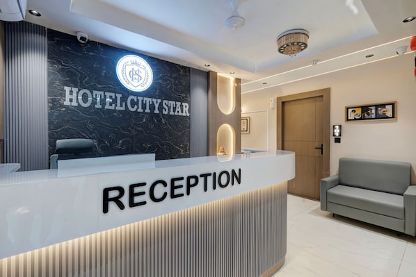 Zona de recepción de hotel con una pared de mármol, mostrador de recepción con la etiqueta 'RECEPTION' y un sofá.
