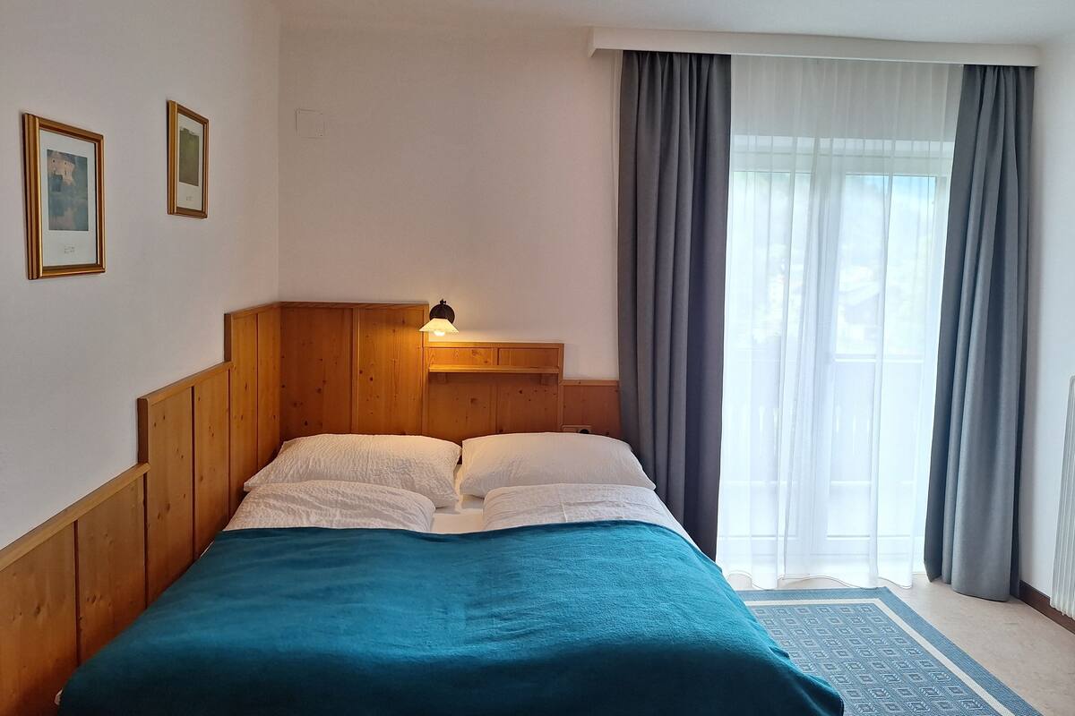 A hotel room with a wooden headboard, a bed with white and teal bedding, a nightstand with a lamp, and a window with curtains.