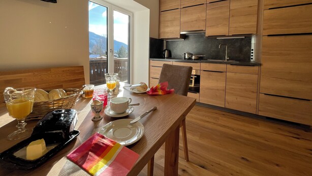 A modern kitchen with wooden cabinets, a dining table set with breakfast items, and a view of mountains through large windows.