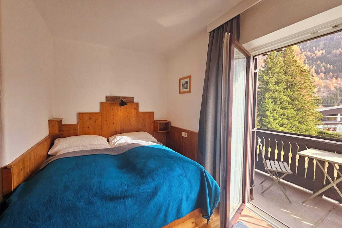 A bedroom with a wooden headboard, a bed with a blue blanket, a nightstand, a picture on the wall, and a balcony with a table and chairs.
