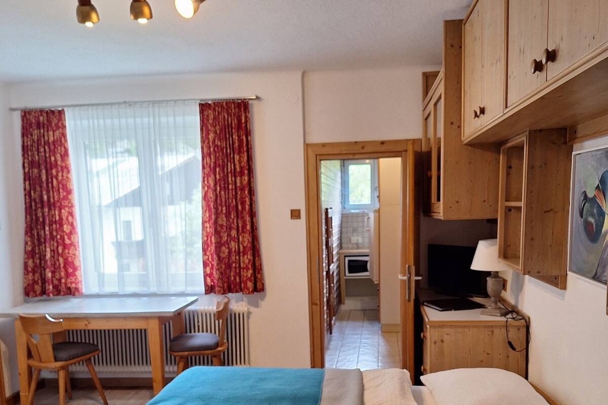 A bedroom with a bed, a desk, and a chair, a kitchenette in the background, and wooden cabinets.