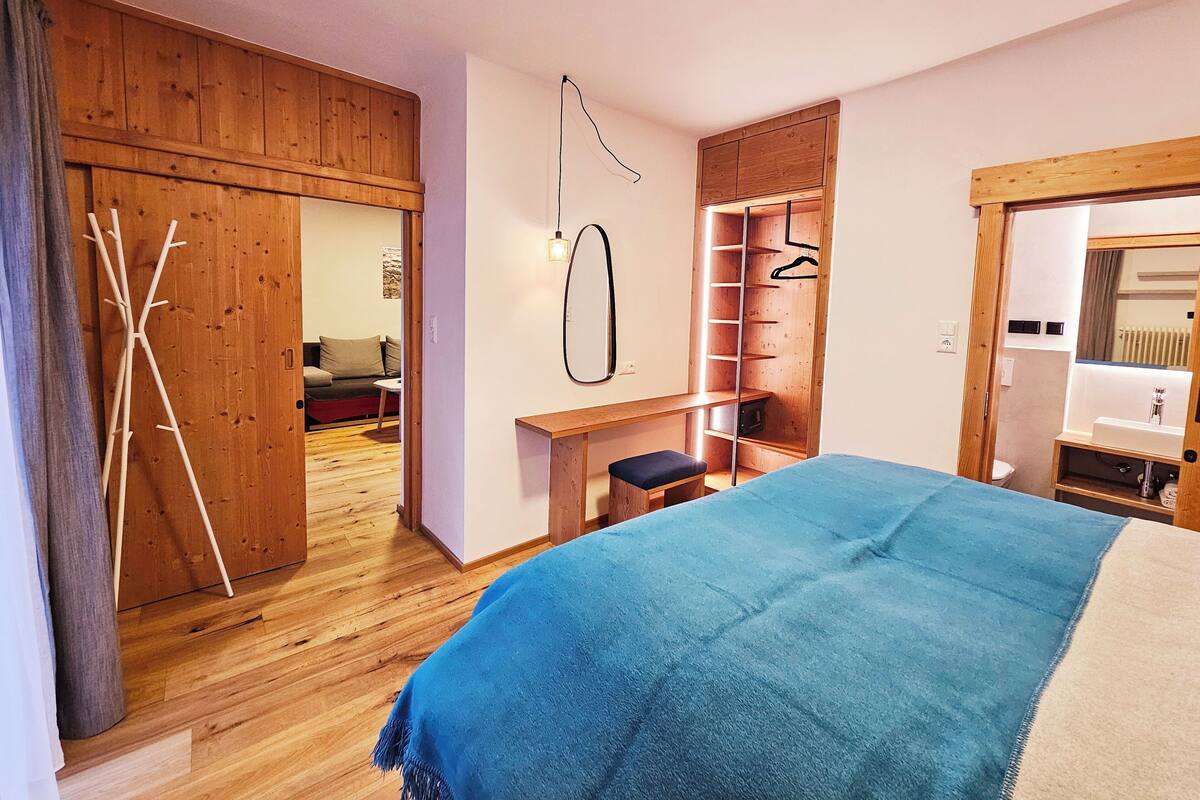 A bedroom with a wooden wardrobe, a bed with a blue blanket, a wooden bench, and a small table.