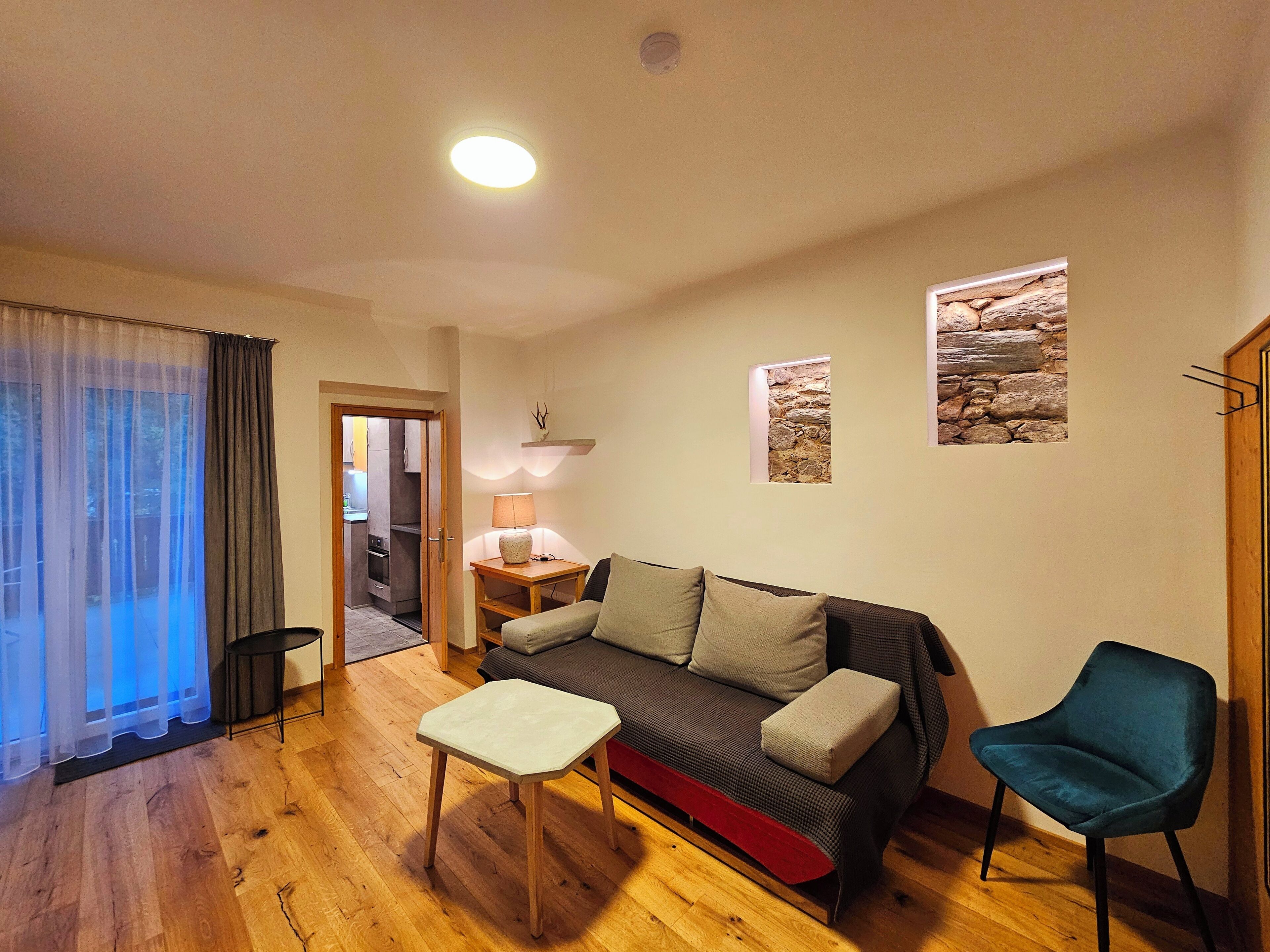 A living room with a sofa, a small table, a chair, and a lamp. There is a door leading to a kitchen and a bathroom.