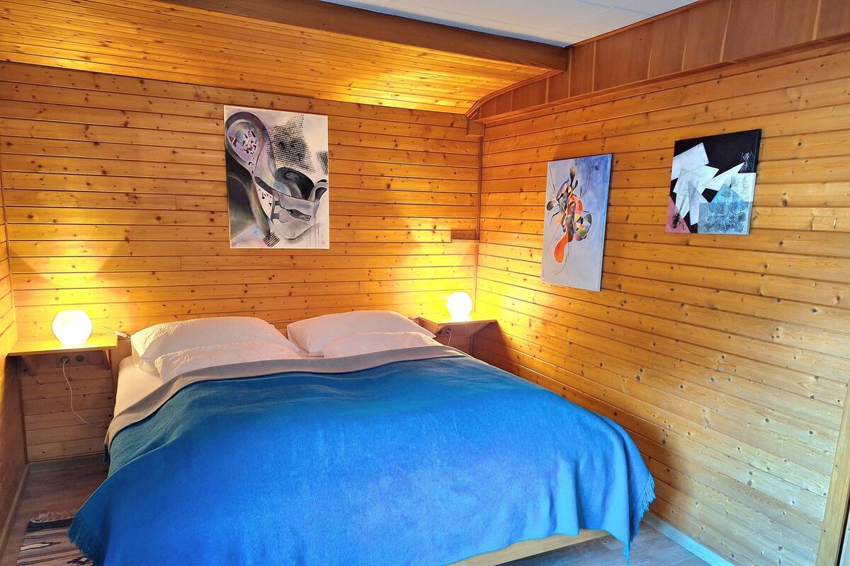 A wooden-paneled room with a bed, two wall-mounted lamps, and three framed artworks.
