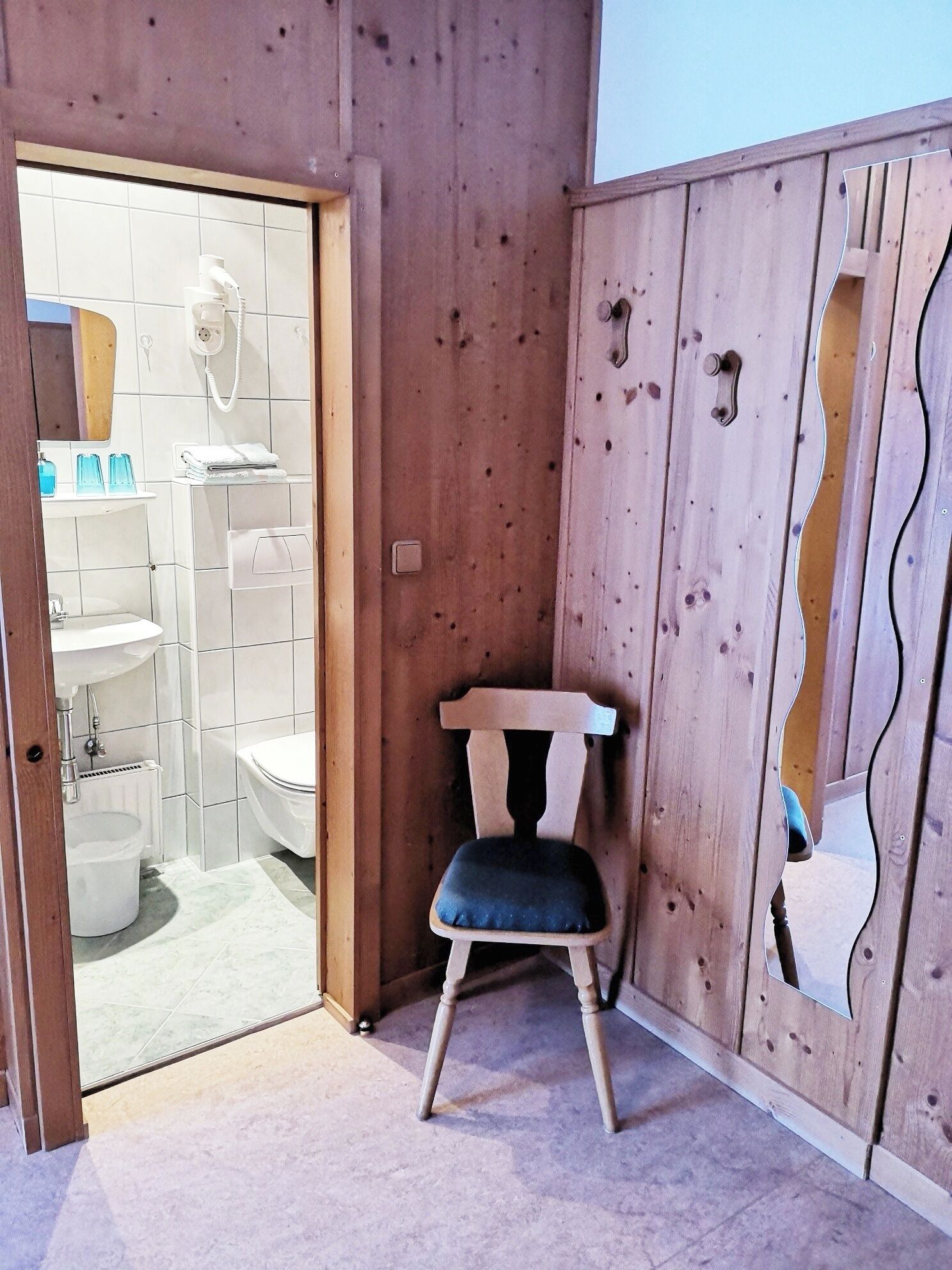 Un intérieur en bois avec une porte en verre donnant sur une salle de bain, une chaise en bois et un mur orné d’un motif original en découpe.