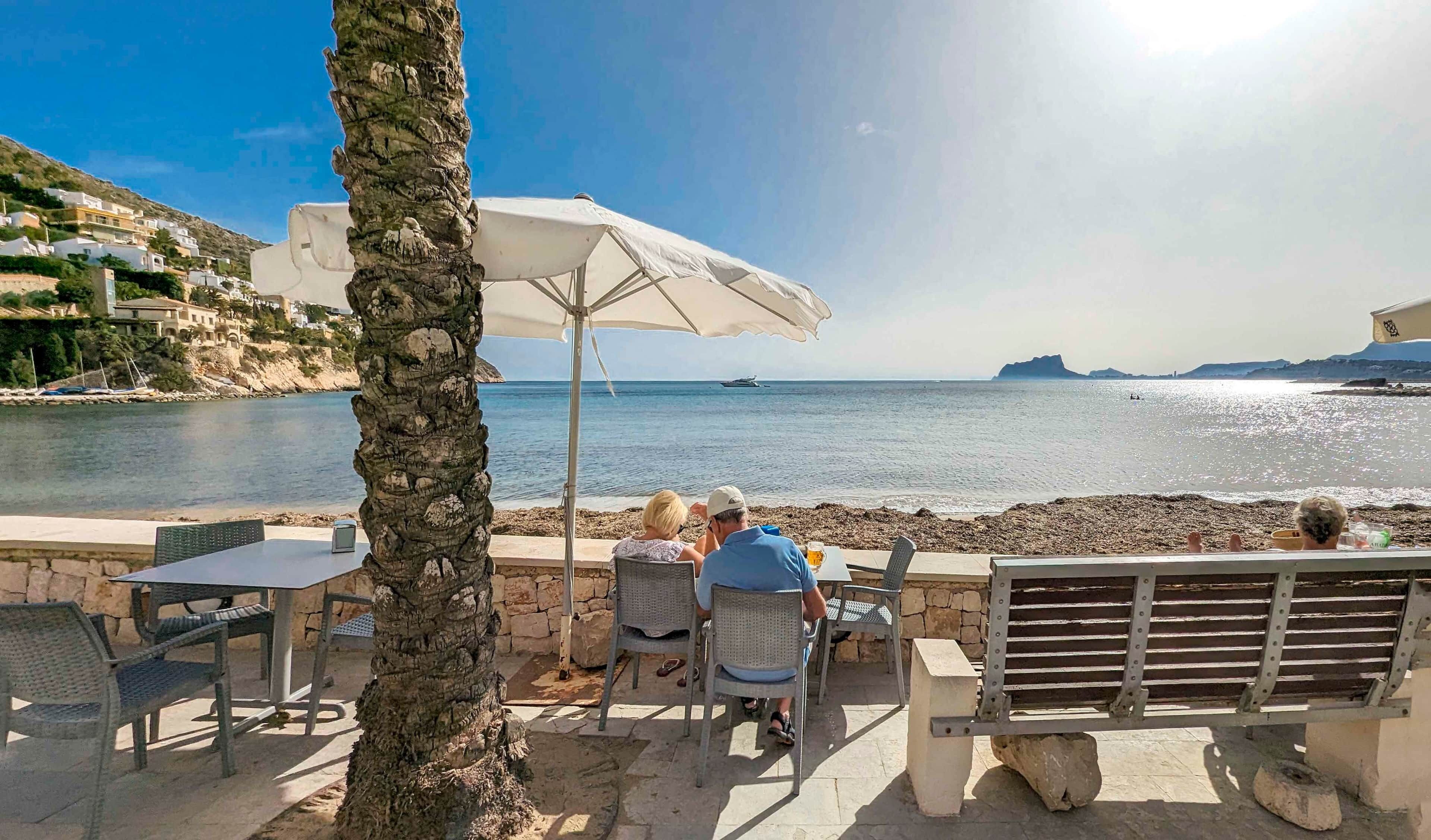 Um terraço à beira-mar com mesa e cadeiras, um grande guarda-sol e vista para o mar e a terra ao longe.