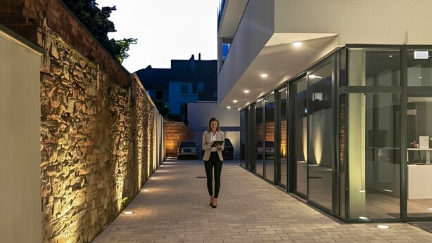 A person walking on a paved pathway between a stone wall and a modern building with glass doors. - GuestHouse Große Linde (Karlsruhe)