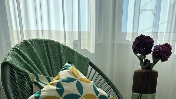 A green chair with a patterned cushion, a small side table with a vase of flowers, and a window with sheer curtains. - GuestHouse Große Linde (Karlsruhe)