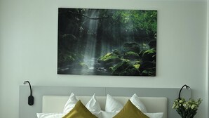 A neatly made bed with white and green linens, a forest-themed wall hanging above, and a bedside table with a vase of flowers. - GuestHouse Große Linde (Karlsruhe)