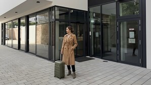 A modern building with large glass doors, a person with a suitcase, and a tiled walkway. - GuestHouse Große Linde (Karlsruhe)
