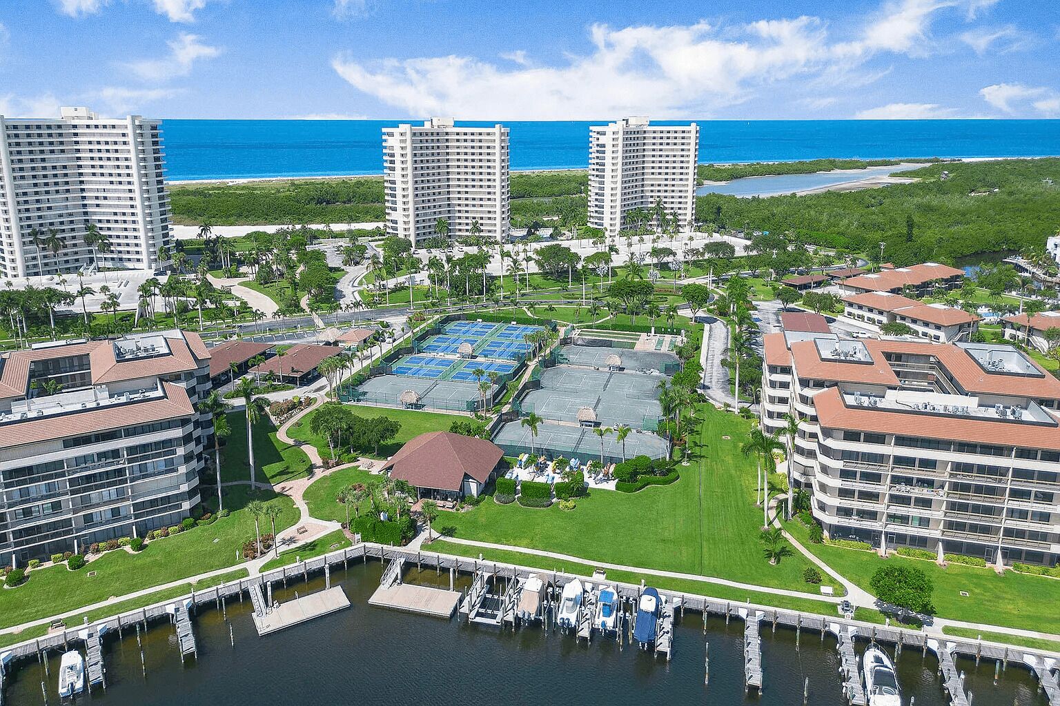Un hotel costero con varios edificios altos, un puerto deportivo con barcos y una cancha de tenis.