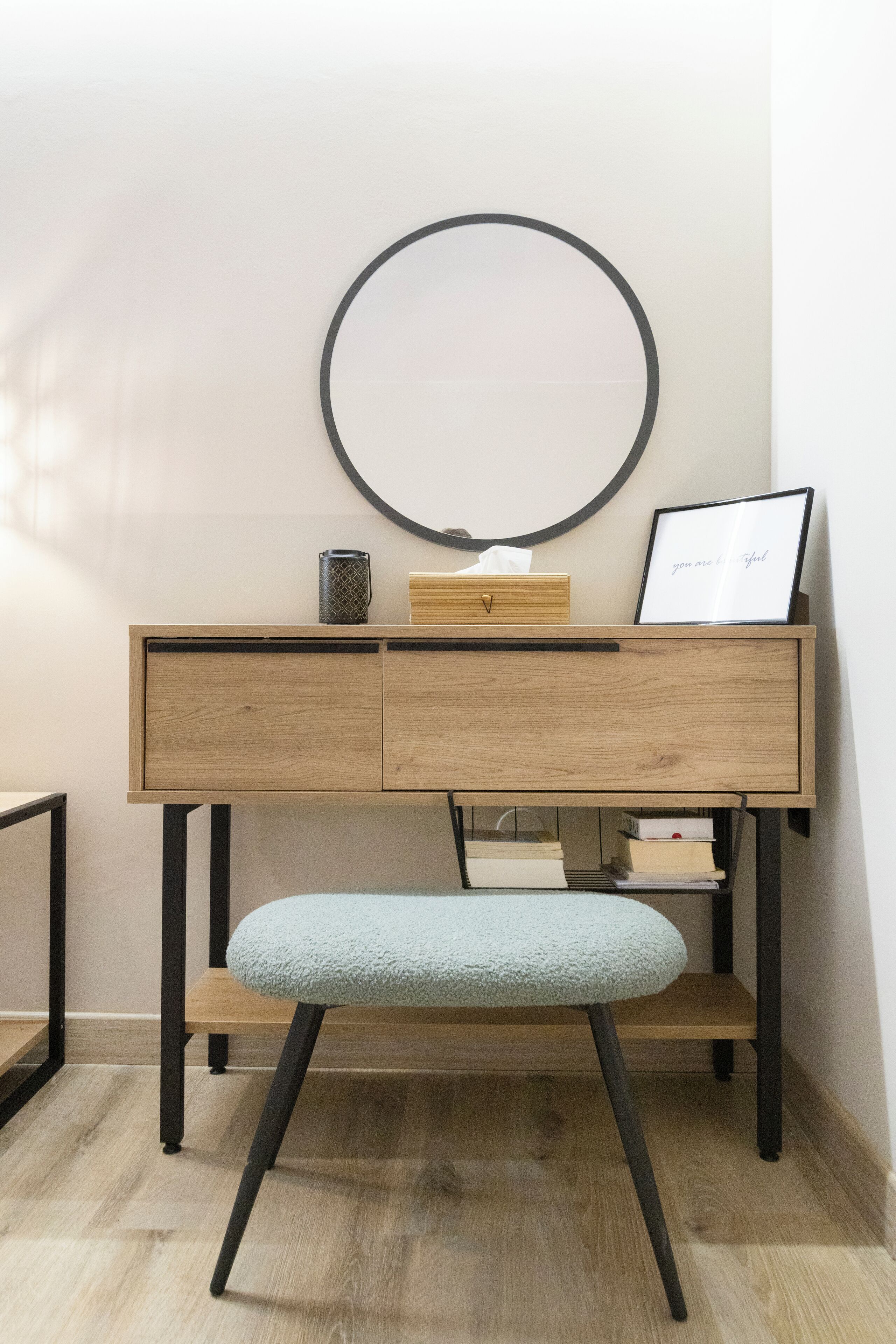 A desk with a light blue cushioned chair, a round mirror, a framed picture, and a small wooden box.
