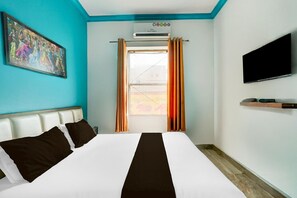 Ett hotellrum med en säng, en tavla på väggen, en luftkonditioneringsanläggning, en tv och ett fönster med gardiner.