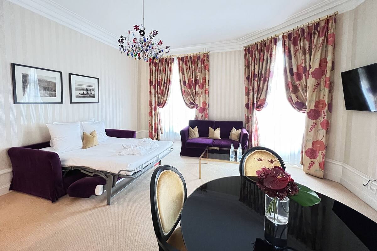 Une chambre d’hôtel comprenant un lit, un canapé, une table à manger, un lustre, des rideaux et un téléviseur à écran plat.