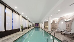 Une longue piscine intérieure, dotée d’un sol carrelé, entourée de murs percés de grandes fenêtres et d’un mur en pierre d’un côté.