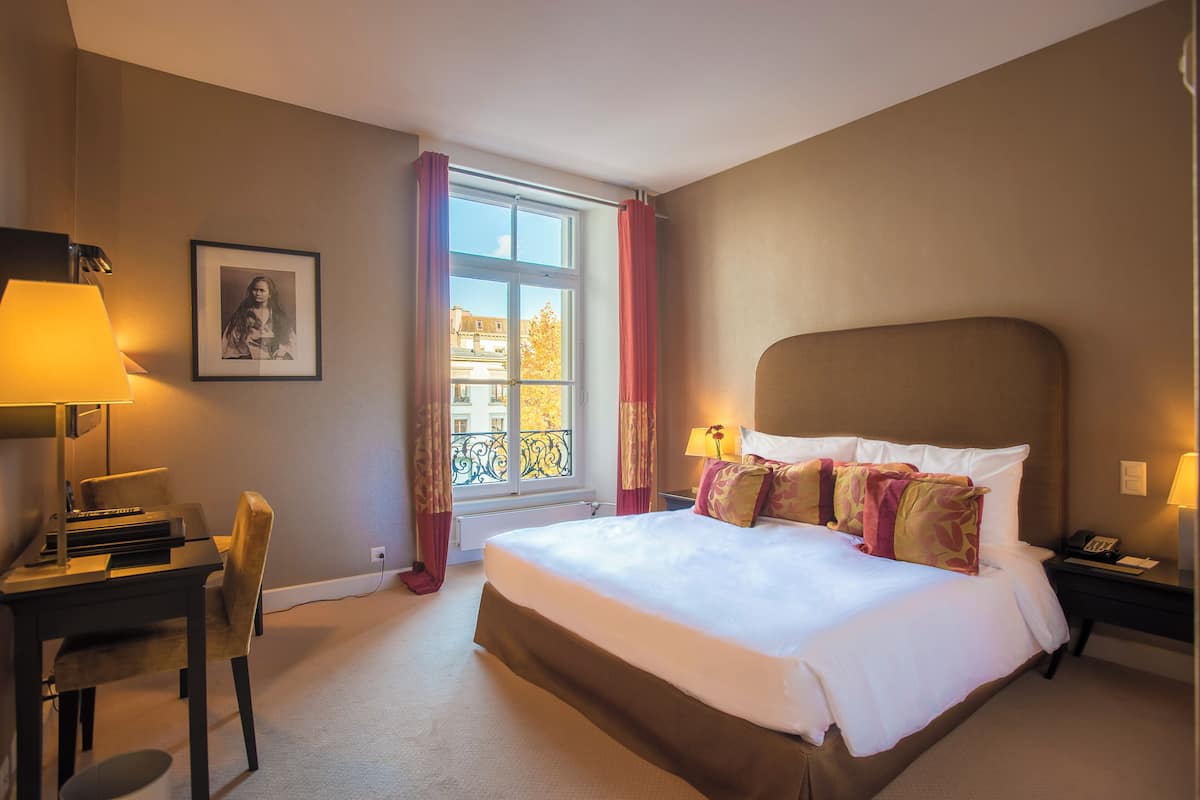 Une chambre d’hôtel avec un lit, un bureau, une chaise, une lampe et une fenêtre avec des rideaux.