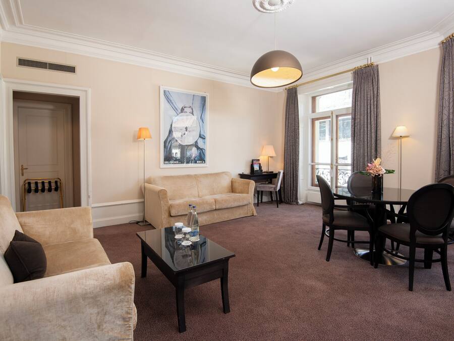 A hotel room with a beige sofa, a glass coffee table, a dining table with chairs, a lamp, and a poster on the wall.