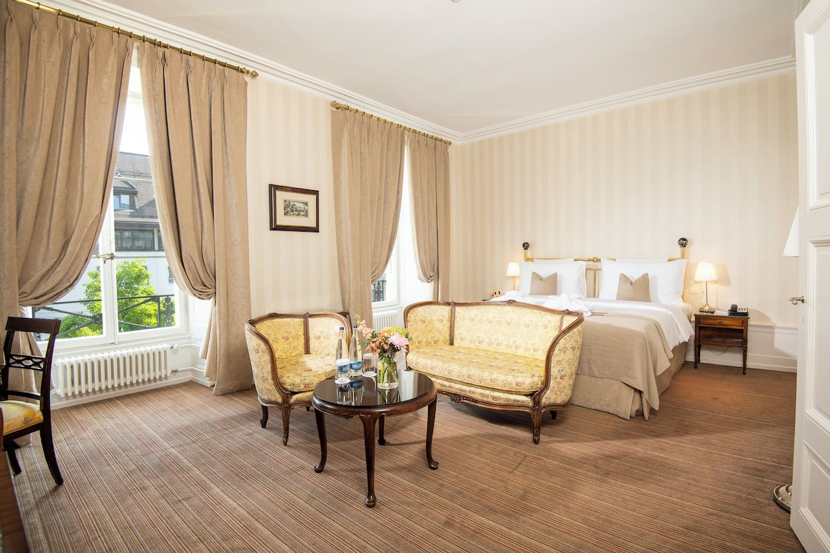 Une chambre d’hôtel avec un canapé, un fauteuil, une table avec un vase, un lit, une table de chevet et une fenêtre avec des rideaux.