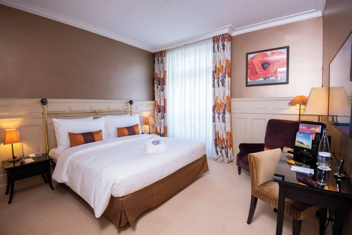 Une chambre d’hôtel avec un grand lit, un bureau avec un ordinateur, une chaise et une fenêtre avec des rideaux.