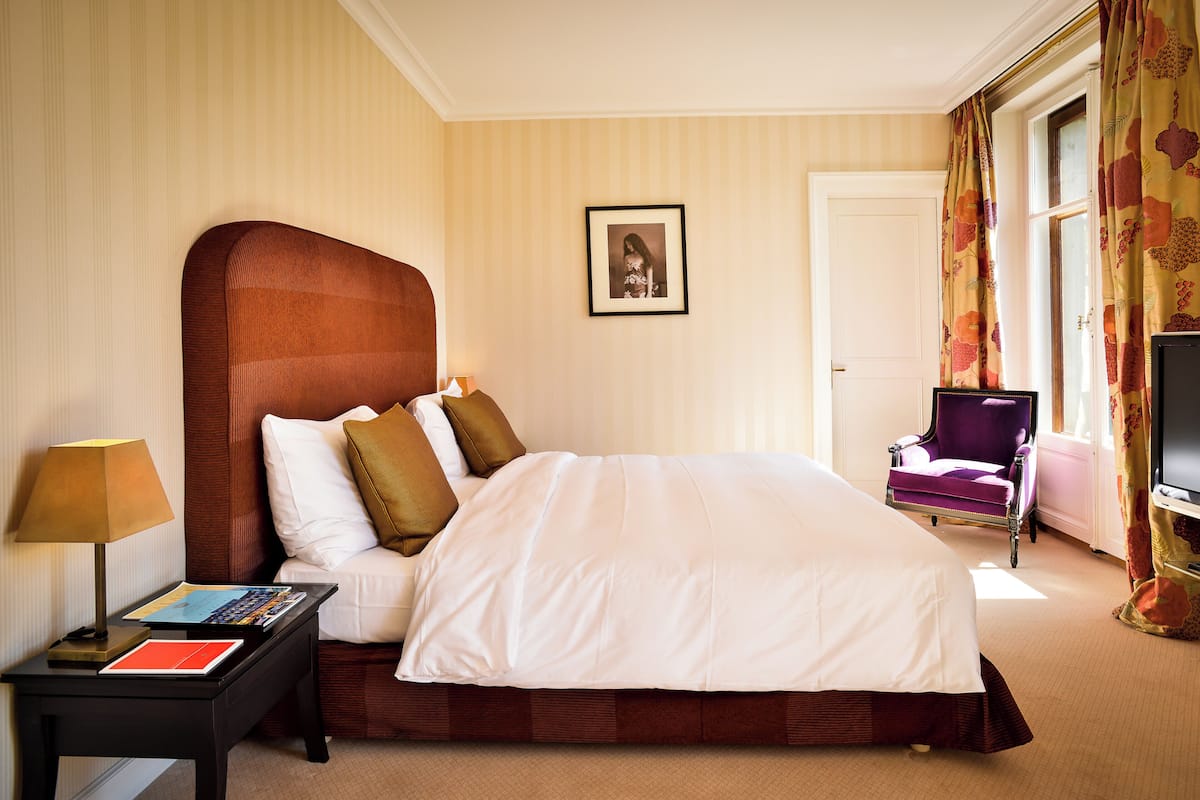Une chambre d’hôtel avec un grand lit, une table de chevet avec une lampe, une chaise, un tableau encadré au mur et un téléviseur.