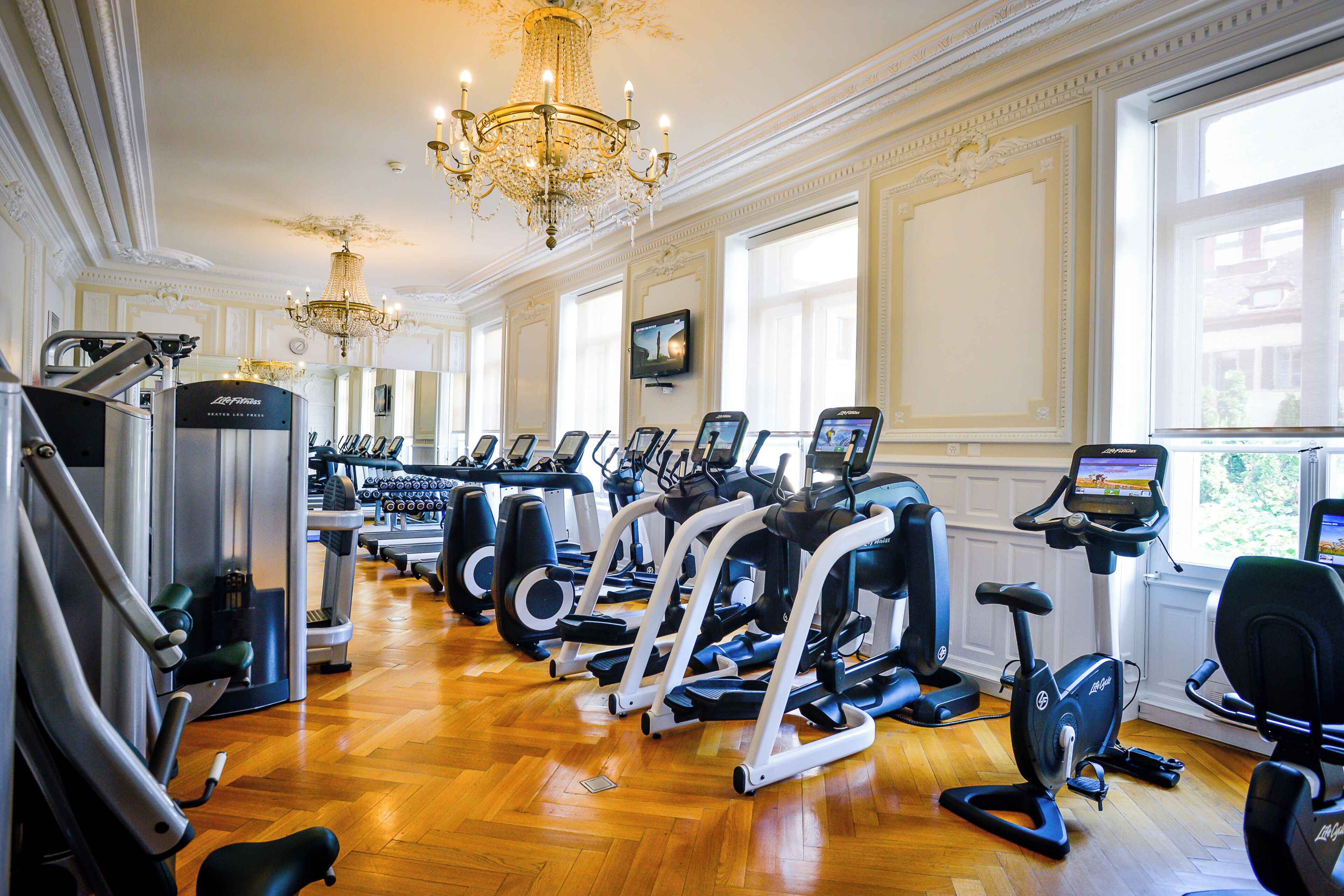 A gym with various exercise machines, a chandelier, and large windows.