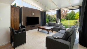A modern living room with a flat-screen TV, a sofa, a coffee table, and a view of a garden through large windows. - Villa Berthelots with Private Pool in Southern Charente (Montmoreau)