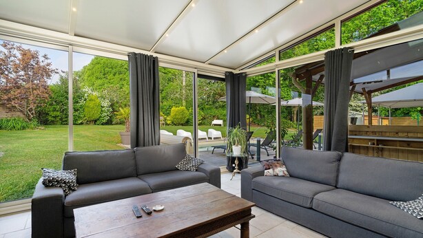 A modern outdoor living area with two grey sofas, a wooden coffee table, and large windows offering a view of a garden and pool. - Villa Berthelots with Private Pool in Southern Charente (Montmoreau)
