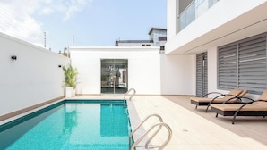 A modern outdoor swimming pool with a glass door entrance, lounge chairs, and a white building with a balcony. - Short Stays By CW (Lagos)