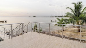 A wooden deck with metal railings overlooking a calm body of water, palm trees, and distant ships. - Short Stays By CW (Lagos)