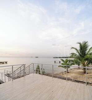 Una terraza de madera con barandas de metal da vista a un cuerpo de agua tranquilo, palmeras y barcos a lo lejos.