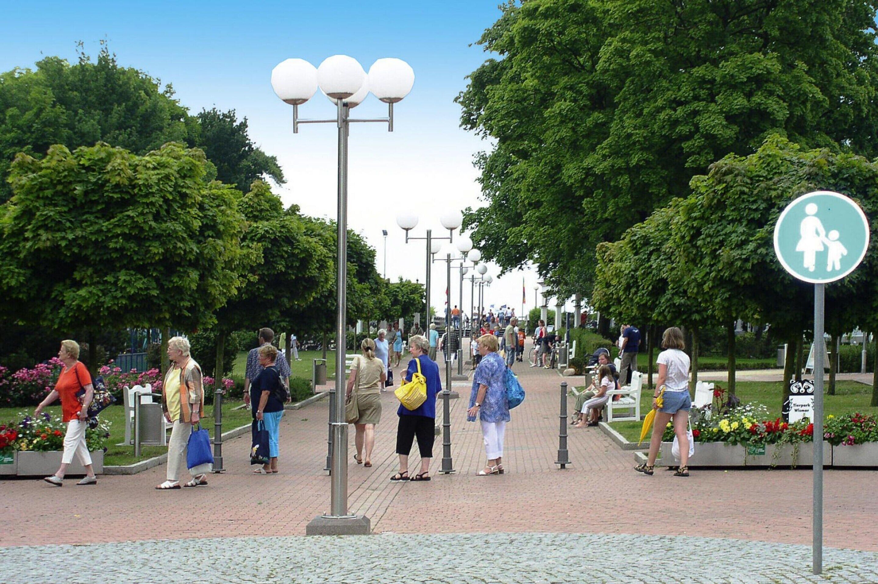 A pedestrian pathway lined with trees and street lamps, with people walking and benches along the sides.