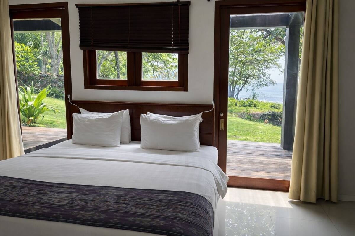 A hotel room with a large bed, two pillows, a headboard, and a view of greenery and water through the windows.