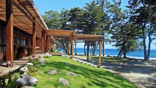 Ein Holzhaus mit einer überdachten Terrasse, einer Rasenfläche mit Steinen und Blick auf das Meer.