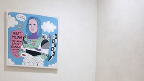 Ein kleiner Raum mit einem hölzernen Hocker, einem Schreibtisch mit Taschentuchbox, einer Fernbedienung und einem Glas. An der Wand hängt ein farbenfrohes Bild.
