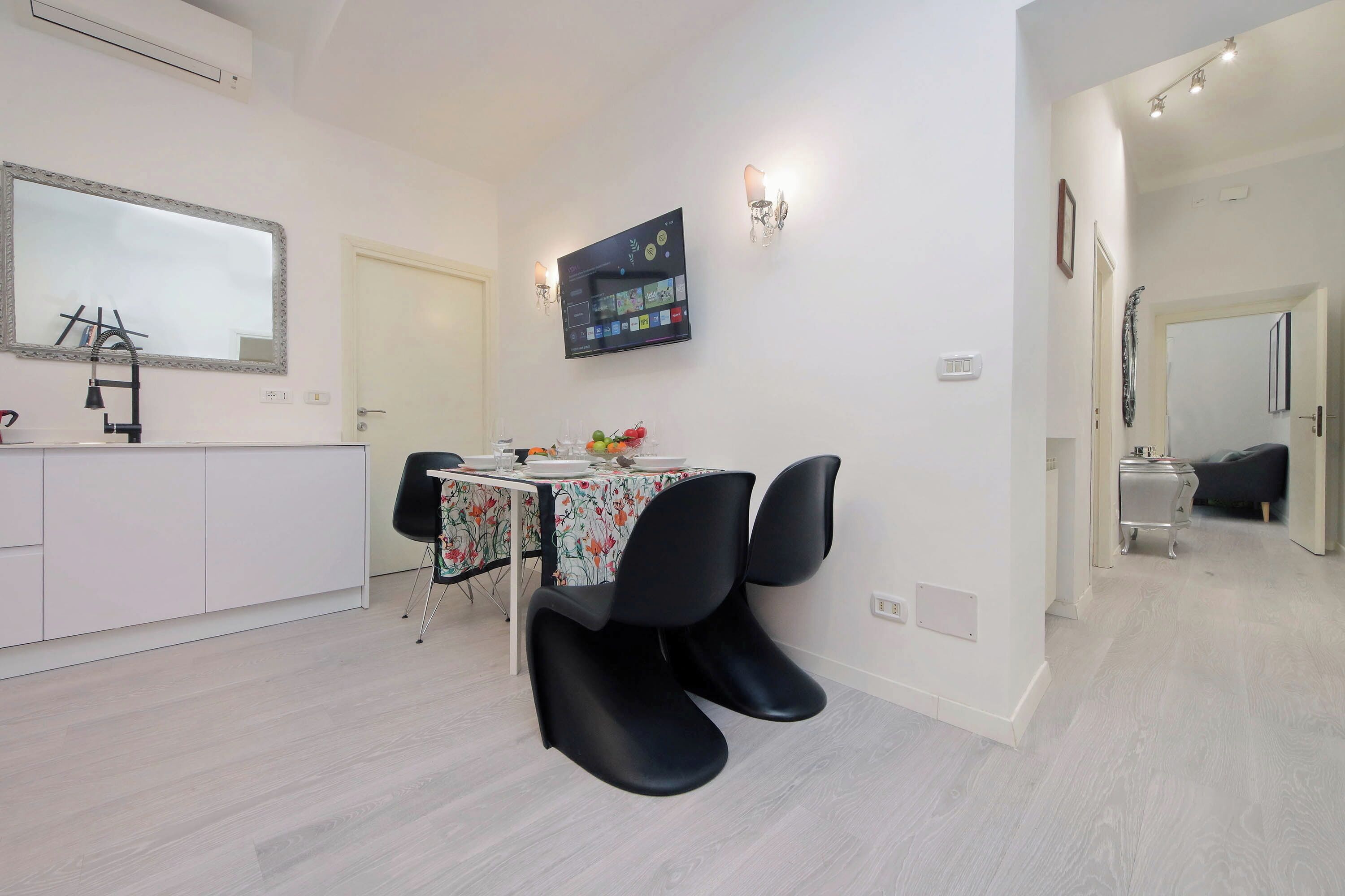 Une cuisine moderne avec un plan de travail blanc, des chaises noires, une table à manger recouverte d’une nappe à motifs floraux et un téléviseur à écran plat fixé au mur.
