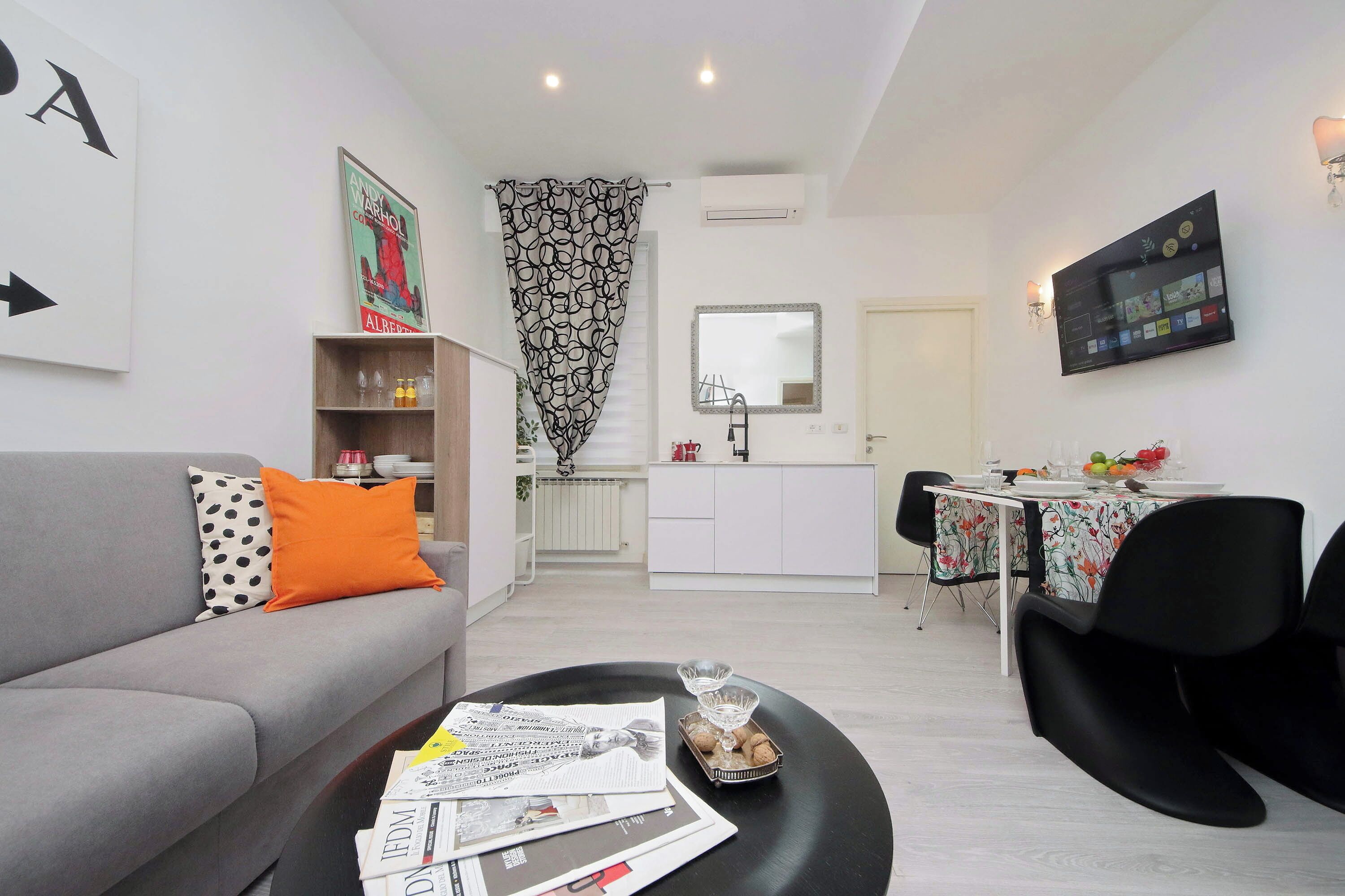 Una sala de estar moderna con un sofá gris, una mesa de centro con periódicos y un comedor con mesa y sillas.