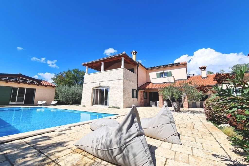 Un área junto a la piscina con sillones reclinables, una piscina y una casa de dos pisos con techo de tejas.