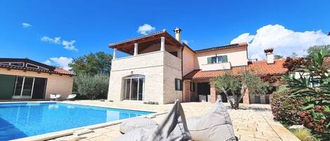 Un área junto a la piscina con sillones reclinables, una piscina y una casa de dos pisos con techo de tejas.