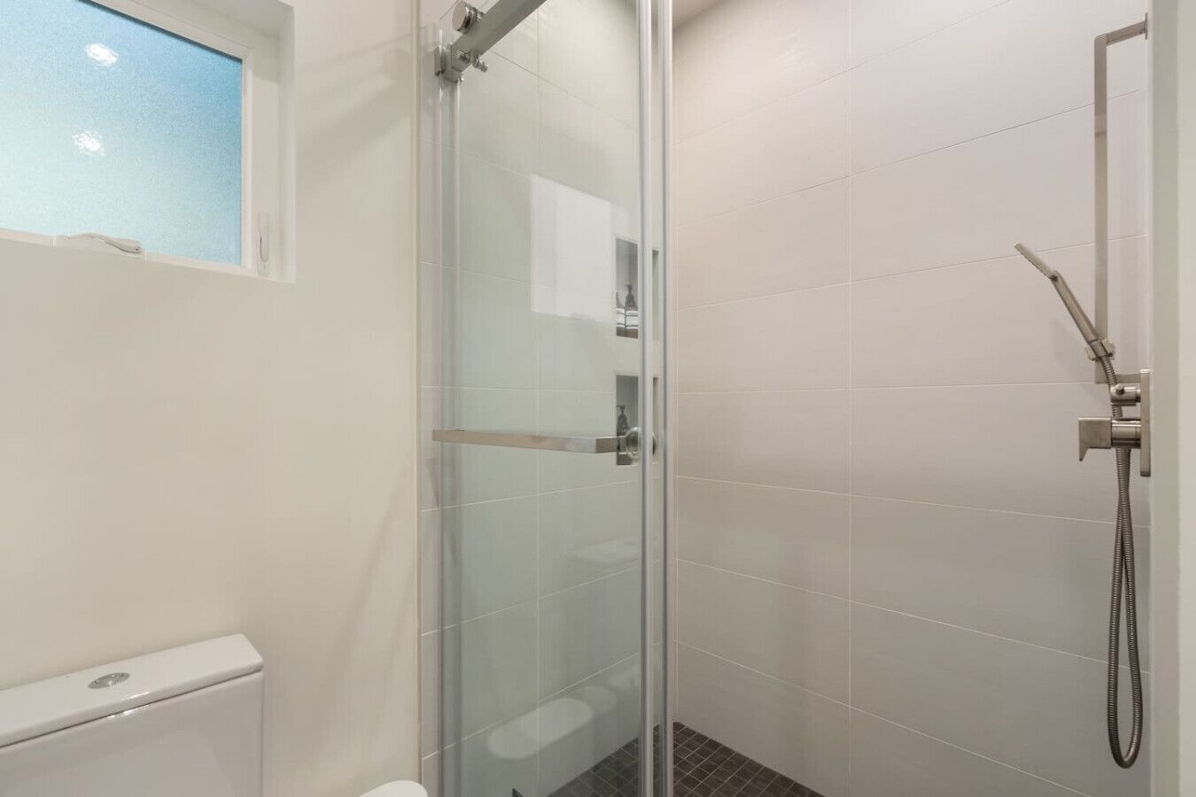 A glass-enclosed shower with a handheld showerhead, a white tiled wall, and a window above the shower.